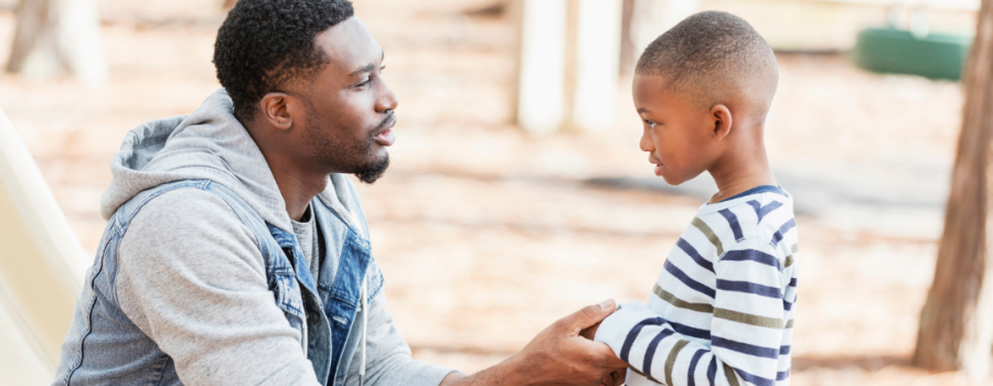 An adult Black male crouching and holding hands with a young Black boy.