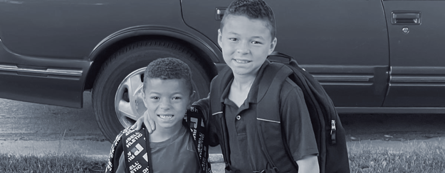 two young boys standing in front of a car with their school backpacks