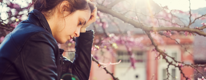 Young woman sitting outside next to a tree with her head in her hand, looking depressed.