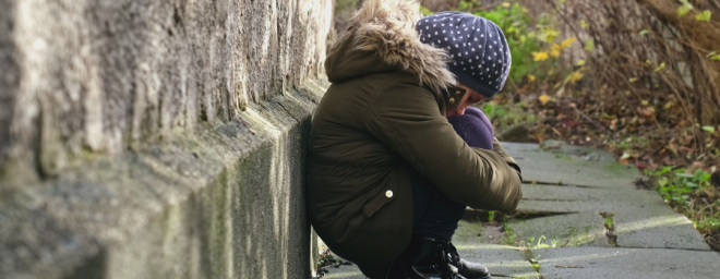 Young person crouching near the ground, holding their knees and looking down.