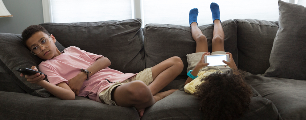 Bored Children sitting on a couch. Teenage boy is holding a TV remote, laying on the couch, while a younger girl is laying upside down on the couch (legs on the backrest) looking at a mobile phone.